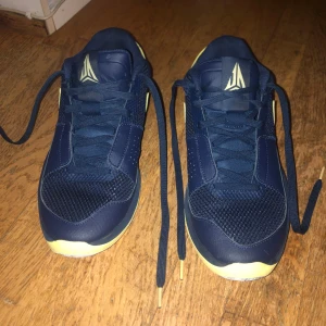 Nike sneakers blå/gul Morant 1 - Snygga Nike Morant 1 sneakers i marinblått med gula detaljer och vit häl. Skorna har mesh och syntet på ovandelen, klassisk snörning och låg profil. Perfekta för dig som gillar sportig stil och vill sticka ut med färg. Swoosh-logga på sidan och nummer 12 broderat bak.