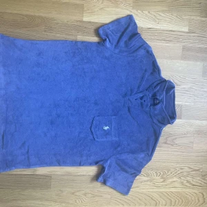 Blå frotté t-shirt från Polo Ralph Lauren - Snygg blå t-shirt i frotté från Polo Ralph Lauren med klassisk krage och bröstficka med broderad logga. Perfekt för sommardagar när du vill vara både chill och stilren. Skön och mjuk känsla tack vare materialet. Storlek M