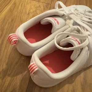 Vita adidas VS Advantage sneakers - Fräscha vita adidas VS Advantage sneakers med rosa detaljer och klassiska ränder baktill. Skorna har snörning, perforerade sidor och rosa detaljer. Aldrig använda, lappen kvar! Hör av er vid frågor!