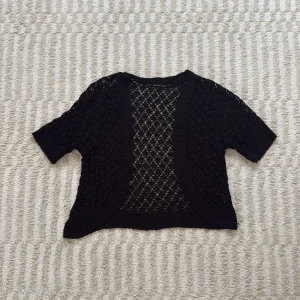 Svart virkad bolero med kort ärm - Svart, kortärmad kofta i virkad stil med öppen front och snyggt hålmönster. Perfekt att slänga över ett linne eller klänning för en chill och trendig look. Koftan är stickad och har ribbade kanter för extra detalj. Den har dock ett hål som går att se på de sista bilderna! Storlek S/M