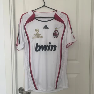 AC Milan Champions League 2007 tröja - AC Milan bortatröja från Champions League-finalen 2007 i Aten. Vit med röda och svarta detaljer, broderat klubbmärke och Adidas-logga. Guldtryck med finalinfo på bröstet och Bwin som sponsor. Tillverkad i lätt och ventilerande polyester.