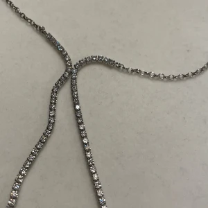 Glittrigt silverhalsband med stenar - Ett elegant halsband i silverfärg med två hängande rader av gnistrande stenar. Kedjan är tunn och smidig, och stenarna ger ett riktigt lyxigt intryck. Perfekt för dig som gillar att sticka ut med lite extra bling.