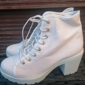 Oanvända Vita boots med klack från DinSko - Snygga vita boots från DinSko i storlek 37. Skorna har kraftig klack, grov sula och snörning framtill. Tillverkade i textil med metallöljetter för en cool look. Perfekta för dig som vill sticka ut med stilrena och trendiga skor.. Ordinariepris 399kr 