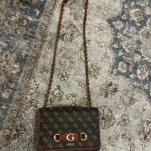 Mönstrad axelväska från Guess - Snygg axelväska från Guess i mörkbrunt skinnliknande material med guldiga detaljer och kedjerem. Väskan har ett klassiskt monogrammönster, rödbruna kanter och en stor G-logga framtill. Baksidan har en praktisk dragkedja för extra förvaring.