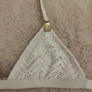 Vit spetsbikini med snäckdetalj - Superfin vit bikini med triangeltopp och matchande trosa, båda i virkad spets med zigzag-mönster. Toppen har en guldig snäckdetalj vid axelbandet och stängs med spänne i ryggen. Aldrig använd och säljer eftersom den är för liten!