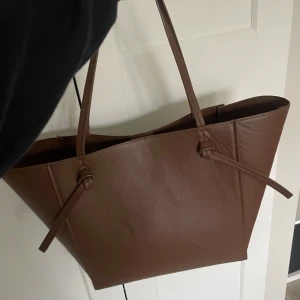 Brun axelväska i skinn med knyt - Stilren brun axelväska i skinn med minimalistisk design och snygga knytdetaljer på sidorna. Väskan är rymlig med långa axelband, perfekt för att bära allt du behöver. Enkel att matcha till olika outfits och passar dig som gillar en clean och klassisk look. Du kan även knyta ihop väskan och göra den mindre för olika tillfällen beroende på vad du behöver😊