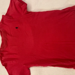 Röd t-shirt från Polo Ralph Lauren - Röd t-shirt från Polo Ralph Lauren med klassisk passform och korta ärmar. T-shirten har en liten mörkblå broderad logga på bröstet och är tillverkad i mjuk bomull. Perfekt för en clean och stilren look.