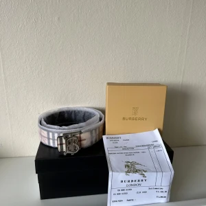 Burberry rutigt skärp med silverdetalj - Snyggt Burberry-bälte med klassiskt rutigt mönster i beige, svart, rött och vitt. Bältet har en stilren silverfärgad 'B'-logga som spänne och är gjort i skinn. Vändbar design med svart sida och diskret Burberry-mönster. Perfekt accessoar för att lyfta din outfit.