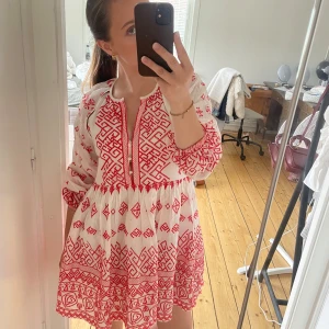 Vit och röd kortklänning med mönster - Superfin vit kortklänning med röda broderade mönster och ballongärmar. Klänningen har v-ringning med knappar framtill och ett luftigt, flowy fall. Perfekt för sommarens alla tillfällen och ger en riktigt bohemisk vibe.