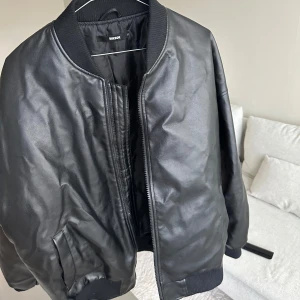 Svart bomberjacka i skinn från Bikbok - Snygg svart bomberjacka i skinn från Bikbok. Jackan har klassisk bombermodell med ribbade muddar vid krage, ärmslut och nederkant. Dragkedja framtill och två fickor. 