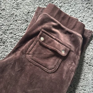 Bruna mjukisbyxor med ficka från juciy couture  - Mysiga bruna mjukisbyxor i mjuk sammet med bred resår i midjan och en stor ficka med lock och tryckknappar på sidan. Perfekta för chill dagar eller när du vill vara extra bekväm.