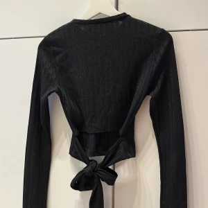 Svart ribbad croppad topp med knyt - Svart långärmad topp med croppad passform. Toppen har knytning i bak:))