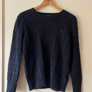 Mörkblå kabelstickad tröja Polo Ralph Lauren - Kabelstickad mörkblå tröja från Polo Ralph Lauren med klassisk rund hals och röd broderad logga på bröstet. Tröjan har långa ärmar och ribbade muddar för en snygg passform. Perfekt för dig som gillar stilren och tidlös design.