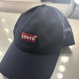 Mörkblå Levi's keps - Snygg mörkblå keps från Levi's med klassisk böjd skärm och röd broderad logotyp framtill. Tillverkad i 100% bomull för en skön och luftig känsla. Den är i bra skick, inga defekter. Vid frågor är det bara att höra av sig!