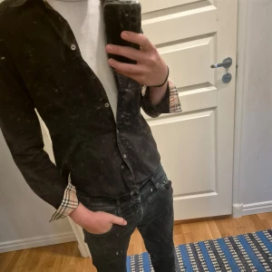 Svart Burberry skjorta  - Säljer en svart skjorta från Burberry i bomull med klassiska rutiga detaljer på insidan av kragen och ärmsluten. Skjortan har knappar framtill och lång ärm. Perfekt för dig som gillar stilrena plagg med en twist. Står M men sitter som S