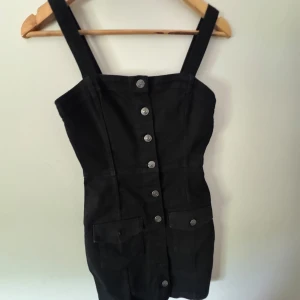 Svart kort klänning med knappar - Snygg svart kort klänning från NLY Trend med breda axelband och knappar framtill. Klänningen har två fickor framtill och är gjord i ett stadigt bomullstyg. Perfekt för dig som gillar en stilren och edgy look.