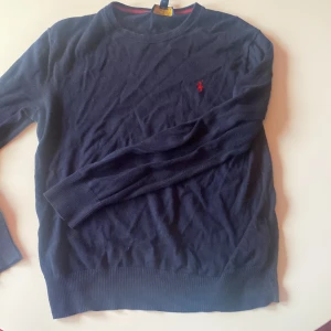 Mörkblå stickad tröja Polo Ralph Lauren - Mörkblå stickad tröja från Polo Ralph Lauren med klassisk röd broderad logga på bröstet. Tröjan har rund halsringning, ribbade muddar och är långärmad. Perfekt för dig som gillar stilren design!