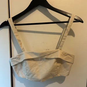 Beige croppad linnetopp - Snygg beige croppad linnetopp med breda axelband och cool detalj framtill. Toppen har en stilren look och dragkedja bak för enkel påklädning. Perfekt att styla med höga jeans eller kjol, passar även bra för semester. Liten i storleken, det är storlek M men känns som S