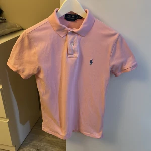 Rosa pikétröja från Polo Ralph Lauren - Säljer en klassisk rosa pikétröja från Polo Ralph Lauren med korta ärmar och blå broderad logga på bröstet. Tröjan har krage och två knappar framtill. Perfekt för en clean och stilren look. Ny tvättad och ostruken på bild endast andvänd fåtal gånger