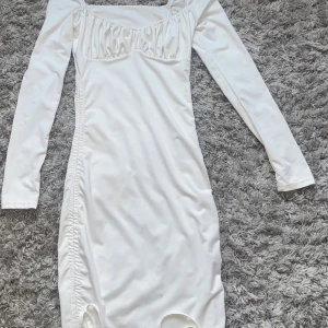 Vit långärmad kortklänning med snörning - Snygg vit kortklänning med långa ärmar och fyrkantig urringning. Klänningen har rynkade detaljer vid bysten och snörning längs ena sidan för en extra cool look. Perfekt för dig som gillar stilrena och trendiga plagg.