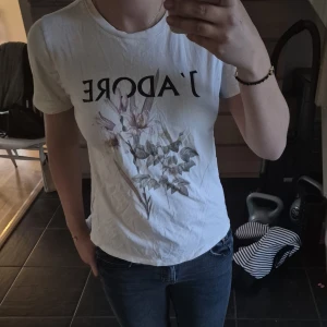 Vit t-shirt med blommor och text - Vit t-shirt med svart texten 'J'ADORE' på bröstet och ett snyggt blommotiv i mjuka färger framtill. Klassisk rund halsringning och korta ärmar. Perfekt för en chill och trendig look.