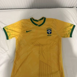 Brasilien fotbollströja Nike gul - Gul Brasilien fotbollströja från Nike med gröna detaljer på krage och ärmslut. Tryckt CBF-emblem och Nike-logga på bröstet samt diskret mönster av Kristusstatyn på både fram- och baksidan. Kortärmad modell i lätt och ventilerande material.