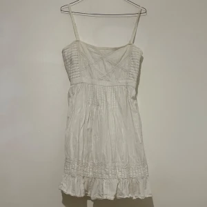 Vit somrig kortklänning med spets - Supersöt vit kortklänning med tunna axelband, volangkant nertill och fina spetsdetaljer på bröstet och längs fållen. Lätt rynkad och luftig modell i bomull, perfekt för varma dagar. Snyggt mönstrad med diskreta ränder och broderade detaljer.