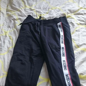 Svarta  moschino dress storlek Mr från Moschino (äkta) - Svarta mjukisbyxor från Moschino med vit och röd logga längs sidorna. Byxorna har snörning i midjan och en bakficka med broderad Moschino-text. Sköna och stilrena, perfekta för chill eller streetwear. Samtidigt så är hoodien skön stilren och bara avslappnad att använda kom dm för fler bilder mvh