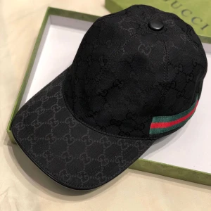 Gucci keps - Snygg svart keps från Gucci med diskret GG-monogram över hela kepsen. Klassisk böjd skärm och ikoniska gröna och röda ränder på sidan. Tillverkad i bomullsmaterial och perfekt för dig som vill ha en stilren och lyxig accessoar.