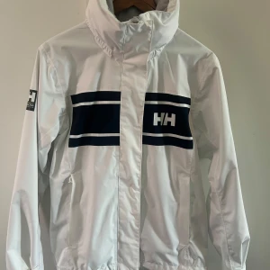 Vit seglarjacka Helly Hansen S - Snygg vit vindjacka från Helly Hansen med marinblå detaljer och logga både fram och bak. Jackan har hög krage, dragkedja och långa ärmar. Perfekt för blåsiga dagar och har en sportig vibe med tydliga ränder över bröstet. Den har en luva som kan vikas ihop. Jackan köptes förra sommaren och endast använder ett fåtal gånger. 
