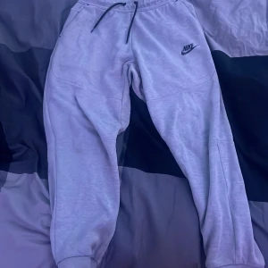 Grå mjukisbyxor från Nike med snörning - Säljer ett par grå mjukisbyxor från Nike i storlek L. Byxorna har svart snörning i midjan, elastiska muddar vid bensluten och en svart Nike-logga på låret. Perfekta för chill eller träning, med mjukt material och dragkedjeficka på ena sidan.