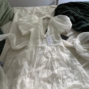 Vit omlottklänning från Gina Tricot - Superfin vit klänning från Gina Tricot med puffärmar, djup v-ringning och stor rosett i midjan. Lätt, krinklat tyg som ger en luftig känsla. Perfekt för sommarens alla tillfällen när du vill känna dig extra söt och trendig.