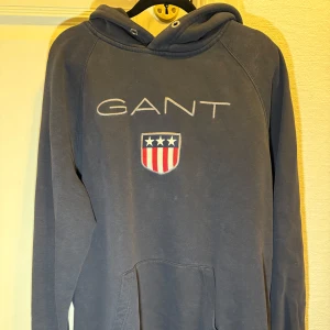 GANT hoodie – stl XXL - Mörkblå hoodie från GANT med broderad logga och sköld. Bekväm och stilren, perfekt till vardags. Använd men i fint skick, inga fläckar eller hål. Snören saknas i luvan.