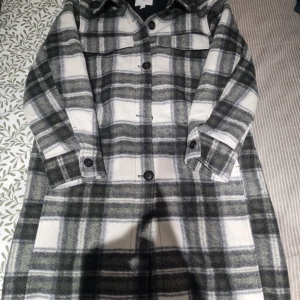 Rutig overshirt i grått och vitt - Snygg lång overshirt med rutigt mönster i grått, vitt och svart. Jackan har stora bröstfickor, knappar framtill och klassisk krage. Perfekt för lager-på-lager och chill stil.