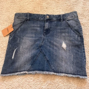 Blå jeanskjol med slitningar - Snygg blå jeanskjol från Sellpy med slitna detaljer och fransad nederkant. Kjolen har klassiska fickor fram och bak och en cool tvättad look. Perfekt för dig som gillar en avslappnad och trendig stil. Oanvänd från Sellpy och kan anpassas i midjan efter önskad storlek (se bild) och passar därför alla storlekar! 