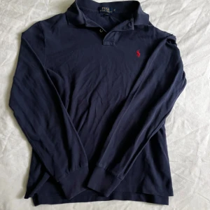 Mörkblå långärmad pikétröja Polo Ralph Lauren - Snygg mörkblå långärmad pikétröja från Polo Ralph Lauren i slim fit. Klassisk krage, knäppning vid halsen och ikonisk röd logga broderad på bröstet. Tillverkad i mjuk bomull, perfekt för en clean och stilren look.