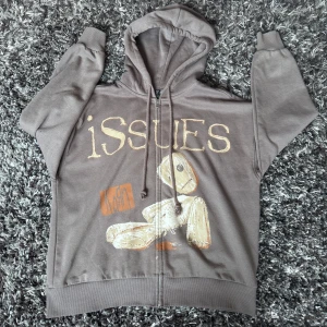 Brun Korn Issues hoodie Divided H&M - Brun hoodie med dragkedja från Divided H&M, storlek S. Framsidan har tryck med Korn Issues-motiv och snörning i huvan. Mjuk bomullsblandning och långa ärmar. Perfekt för dig som gillar bandmerch och streetstyle.