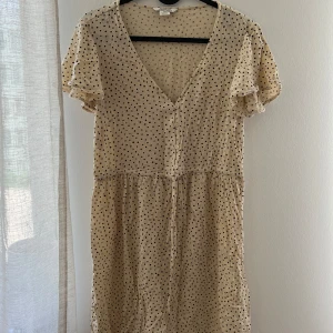 Klänning från Monki - Supersöt beige kortklänning från Monki med svarta prickar, v-ringning och knappar framtill. Klänningen har korta, lite fladdriga ärmar och ett lätt, luftigt tyg som passar perfekt till varma dagar. 
