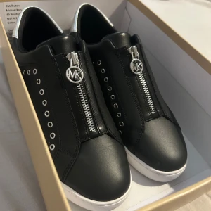 Svarta sneakers från Michael Kors - Använda en gång. Stilrena svarta sneakers i skinn från Michael Kors med vita sulor och silvriga detaljer. Dragkedja framtill med MK-logga. 