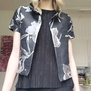 Blommig väst/kofta från Herno - Snygg svart väst från Herno med stora grå och vita blommor. Jackan har korta ärmar, krage och guldiga knappar framtill. Är öppen för prisförslag!💕 Köpt för 4690