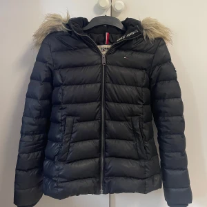 Svart pufferjacka Tommy Jeans - Snygg svart pufferjacka från Tommy Jeans med huva och fuskpälsdetalj. Jackan har dragkedja framtill, två fickor och klassisk quiltad design. Perfekt för kalla dagar och riktigt skön att bära. Stilsäker och enkel att matcha med allt. Mycket fint skick enda defekterna är de små märkena på undersidan av armarna (se bilder). 