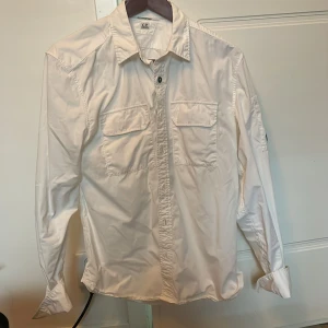 Vit overshirt från C.P. Company - Säljer en stilren vit overshirt från C.P. Company med klassisk krage, två bröstfickor med lock och ikonisk linsdetalj på ärmen. Skjortan har långa ärmar och knäppning framtill. Perfekt för dig som gillar streetwear och snygga detaljer. Nypris runt 2700kr. En knapp som gått sönder därav priset, dm för bild. 🙌🍺