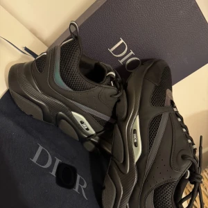 Svarta sneakers  - Chunky svarta sneakers från Dior med meshpaneler och detaljer i skinn och syntet. Snygga grå och mörka inslag, samt tydlig Dior-logga på sidan. Skorna har snörning och en kraftig platt sula för en modern look.