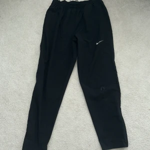 Svarta mjukisbyxor från Nike - Svarta mjukisbyxor från Nike med klassisk logga på låret. Byxorna har resår och snörning i midjan för skön passform. Perfekta för chill eller träning, tillverkade i mjukt material som känns bekvämt hela dagen. 