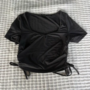 Svart croppad topp med knyt från SHEIN - Svart croppad topp från SHEIN i stretchigt material. Toppen har korta ärmar, v-ringning och snygga knytband i sidorna som ger en trendig look. Perfekt att matcha med höga jeans eller kjol för en cool stil.