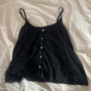 Svart linnetopp med knappar - Svart linnetopp med smala justerbara axelband och vita knappar framtill. Toppen har en lös passform och v-ringning, perfekt för varma dagar. Enkel och stilren design som funkar till många olika outfits.