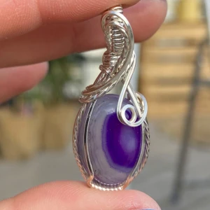 Silverberlock med lila agat - Unikt halsband med en oval lila agat infattad i intrikat lindad silvertråd. Stenen har vackra skiftningar i lila och grått, och infattningen har snygga snirkliga detaljer som ger ett handgjort och konstnärligt intryck. Perfekt statement piece för dig som gillar färg och unika smycken.