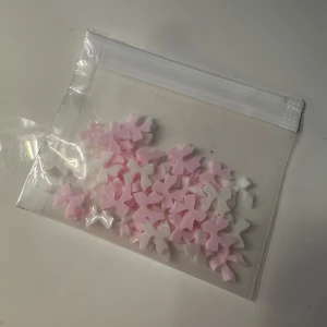 Söta rosa och vita rosetter till naglar - Ett gäng små dekorativa rosetter i rosa och vitt, perfekta för att pynta naglarna och skapa en gullig look. Kommer i en smidig zip-påse. Materialet är plast och rosetterna är formade för att enkelt fästas på naglarna med nagellim.