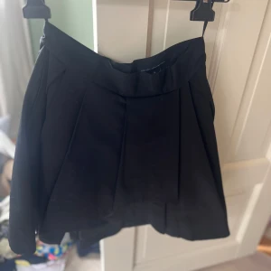 Svart kortkjol från French Connection - Svart kortkjol från French Connection med snygga veck och hög midja. Kjolen har en klassisk siluett och stängs med knapp och dragkedja fram. Perfekt för dig som gillar stilrena och enkla plagg med en modern touch.