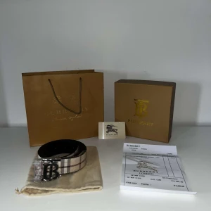 Burberry rutig skärp med B-spänne - Snyggt Burberry-bälte i klassiskt rutigt mönster med beige, bruna och svarta toner. Bältet har ett stort metallspänne med Burberrys ikoniska B-logga. Tillverkat i kalvskinn och levereras med originalpåse, box och kvitto.